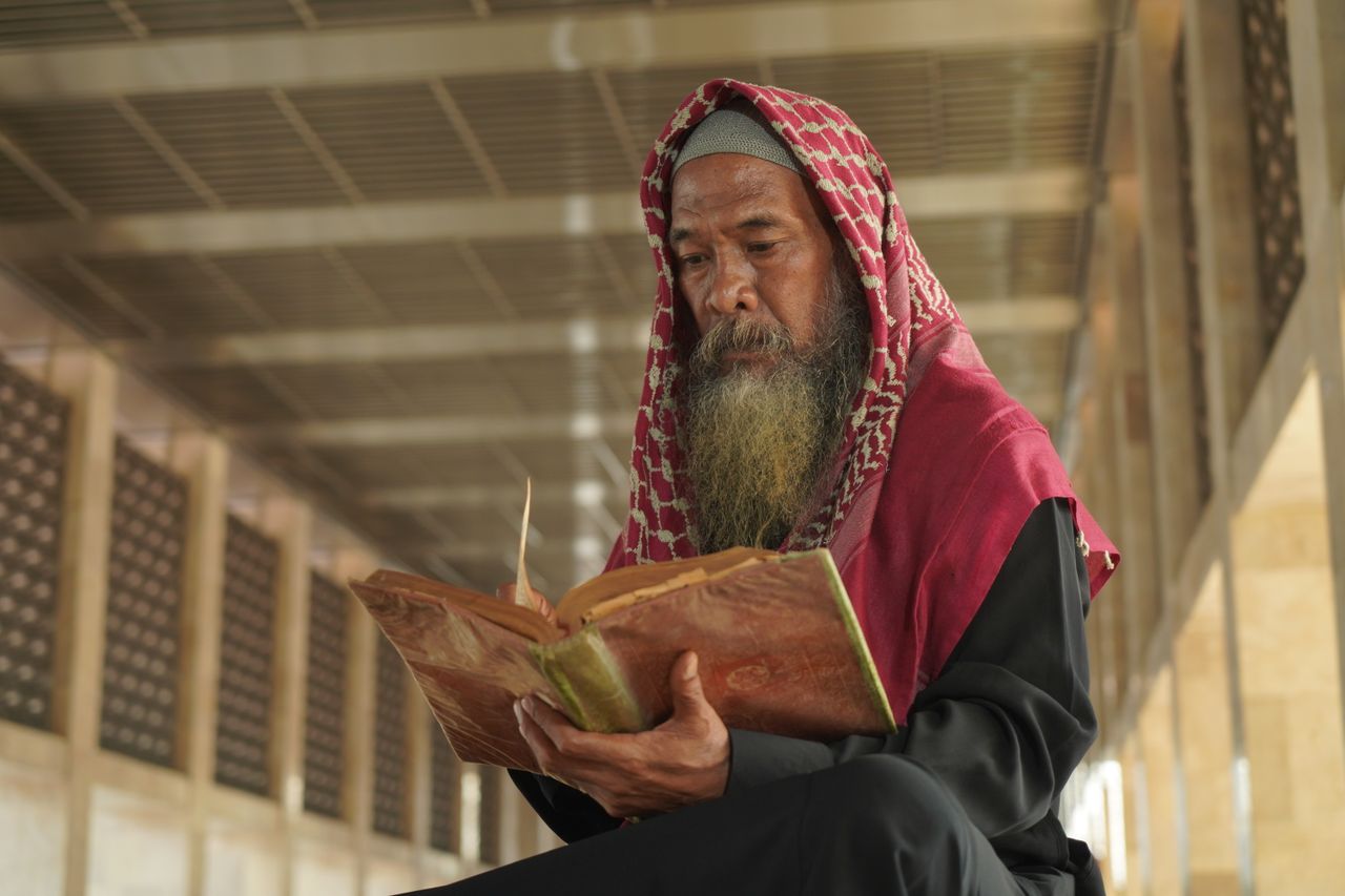 Services Elderly man in traditional attire reading a book indoors, capturing cultural richness in Jakarta.